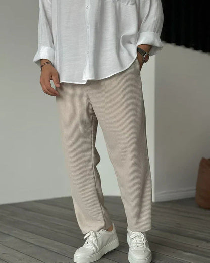 Weiße Hemdbluse und beige, gerippte Hose kombiniert mit weißen Sneakern auf Holzboden. Modetrend, lässiger Stil, Herrenmode, minimalistisch.