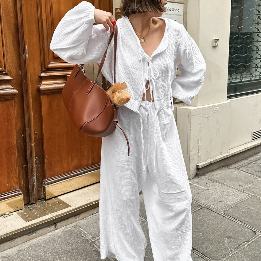 Frau in weißem Leinen-Outfit mit brauner Ledertasche und Baguette, steht vor Holztür. Streetstyle, Sommermode, lässiger Look.