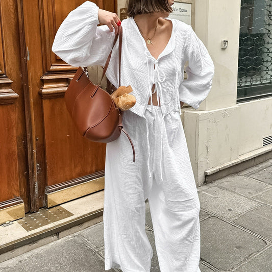 Frau in weißem Leinen-Outfit mit brauner Ledertasche und Baguette, steht vor Holztür. Streetstyle, Sommermode, lässiger Look.