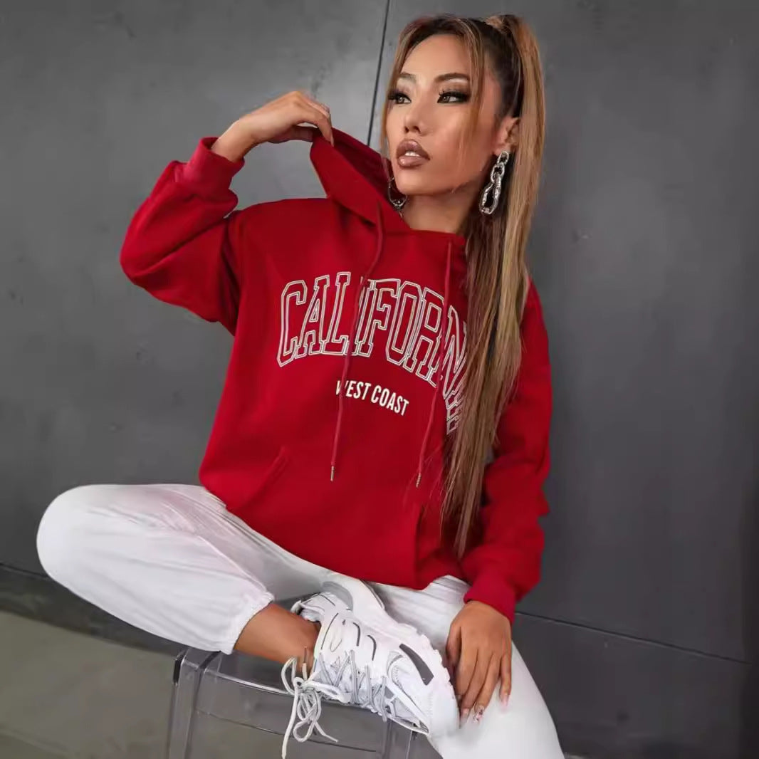 Frau in rotem "California West Coast" Hoodie, weiße Hose und Sneakers, sitzt lässig auf einem transparenten Stuhl vor grauer Wand. Streetwear, Mode.