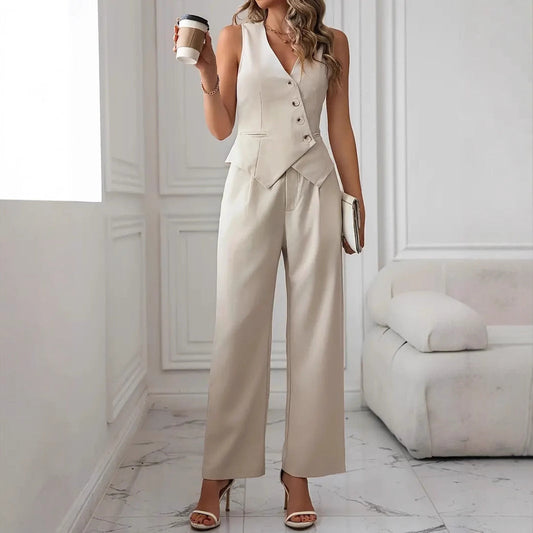 Frau in eleganter, cremefarbener Weste und Hose, hält Kaffeebecher, steht in modernem, minimalistischem Wohnzimmer mit weißem Sofa.