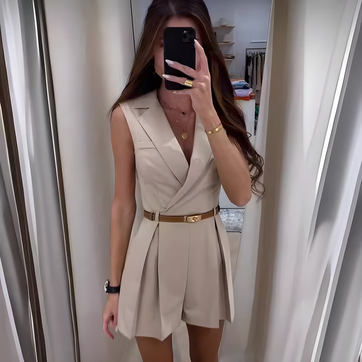 Frau in beigem ärmellosen Jumpsuit mit Gürtel, Spiegel-Selfie, modisches Outfit, elegantes Design, lange Haare, goldene Accessoires.