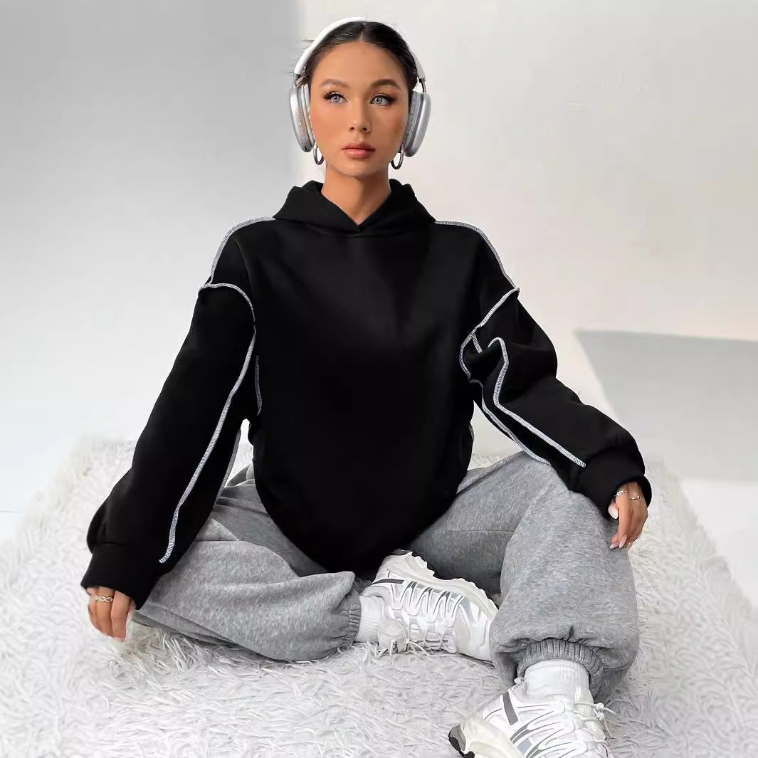 Frau mit Kopfhörern in schwarzem Hoodie und grauer Jogginghose sitzt auf Teppich, trägt weiße Sneaker. Entspannte Mode, Streetwear, Musik hören.