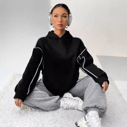 Frau mit Kopfhörern in schwarzem Hoodie und grauer Jogginghose sitzt auf Teppich, trägt weiße Sneaker. Entspannte Mode, Streetwear, Musik hören.