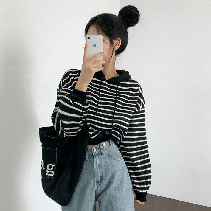 Frau in gestreiftem Hoodie und blauer Jeans macht Selfie mit Smartphone, trägt schwarze Tasche. Modetrend, lässiger Stil, Streetwear-Look.