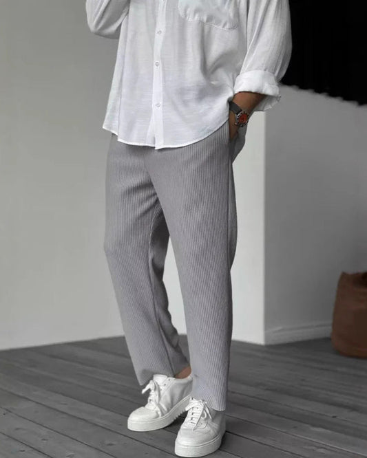 Mann in weißem Hemd und grauer Hose, trägt weiße Sneaker. Moderne Herrenmode, lässiger Stil, Holzfußboden, minimalistische Einrichtung.