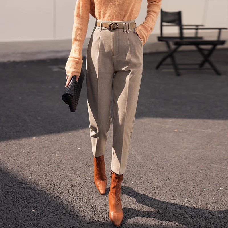 Dame in eleganter, beiger Hose und braunen Stiefeln, trägt orangefarbenen Pullover und schwarze Clutch, modisch gekleidet auf Asphalt.