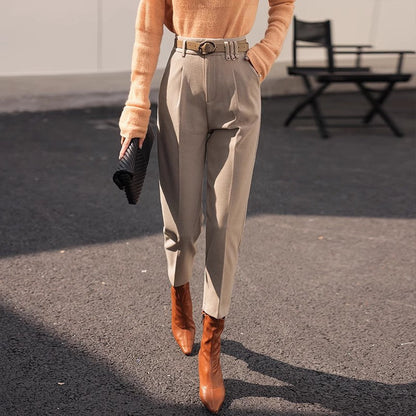 Dame in eleganter, beiger Hose und braunen Stiefeln, trägt orangefarbenen Pullover und schwarze Clutch, modisch gekleidet auf Asphalt.