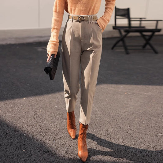Dame in eleganter, beiger Hose und braunen Stiefeln, trägt orangefarbenen Pullover und schwarze Clutch, modisch gekleidet auf Asphalt.