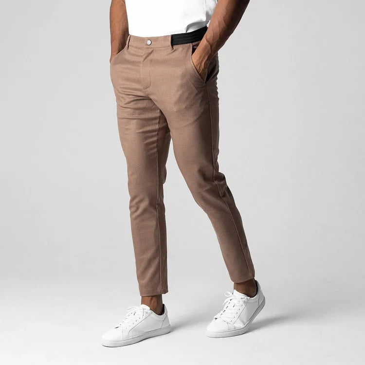 Mann trägt beige Chinohose und weiße Sneaker, Hände in den Taschen, vor grauem Hintergrund. Modische Herrenbekleidung, lässiger Stil.