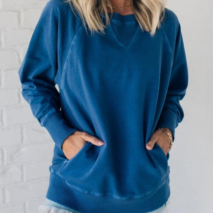 Frau in blauem Sweatshirt mit langen Ärmeln, lässiger Stil, Hände in Taschen, vor weißer Ziegelwand. Bequemes Freizeitoutfit.