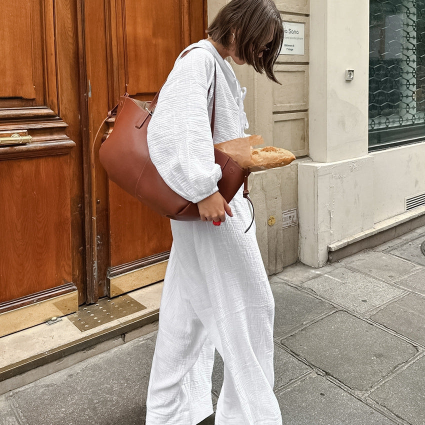 Frau in weißem Jumpsuit trägt braune Ledertasche mit Baguette auf Pariser Straße. Mode, Einkauf, Paris, Baguette, Ledertasche.