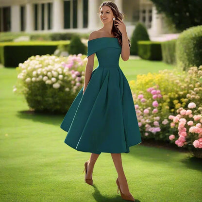 Frau in elegantem, schulterfreiem, smaragdgrünem Kleid auf gepflegtem Rasen mit blühenden Sträuchern im Hintergrund, Sommermode, Gartenparty.