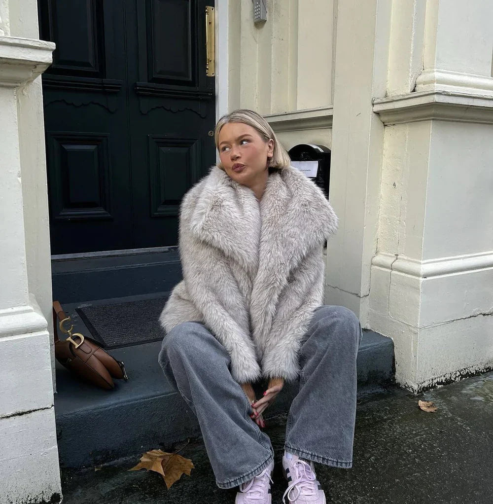Frau in grauem Kunstpelzmantel sitzt auf Treppe vor schwarzer Tür, trägt weite Jeans und weiße Sneaker. Herbstlaub auf dem Boden. Urbaner Stil.
