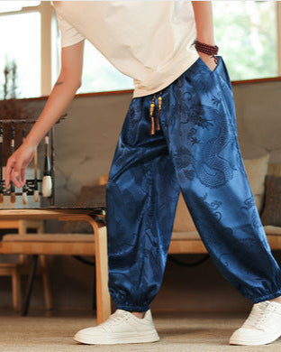Person in blauer Seidenhose mit Drachenmuster, weißem T-Shirt und weißen Sneakers in modernem Wohnzimmer. Asiatische Mode, lässiger Stil.