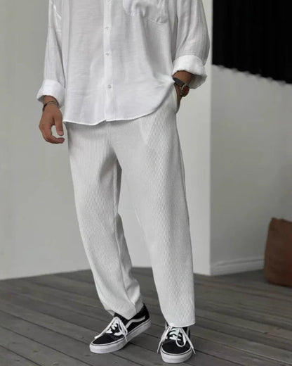 Mann in weißem Hemd und weißen Hosen, trägt schwarze Vans-Sneaker. Moderner, minimalistischer Stil, lässige Herrenmode, Holzboden.
