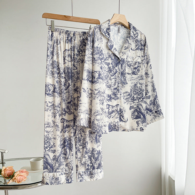 Zweiteiliger Pyjama-Set aus Seide mit blau-weißem Toile-de-Jouy-Muster, hängend auf Holzkleiderbügeln, neben einem Tisch mit Rosen und Tasse.