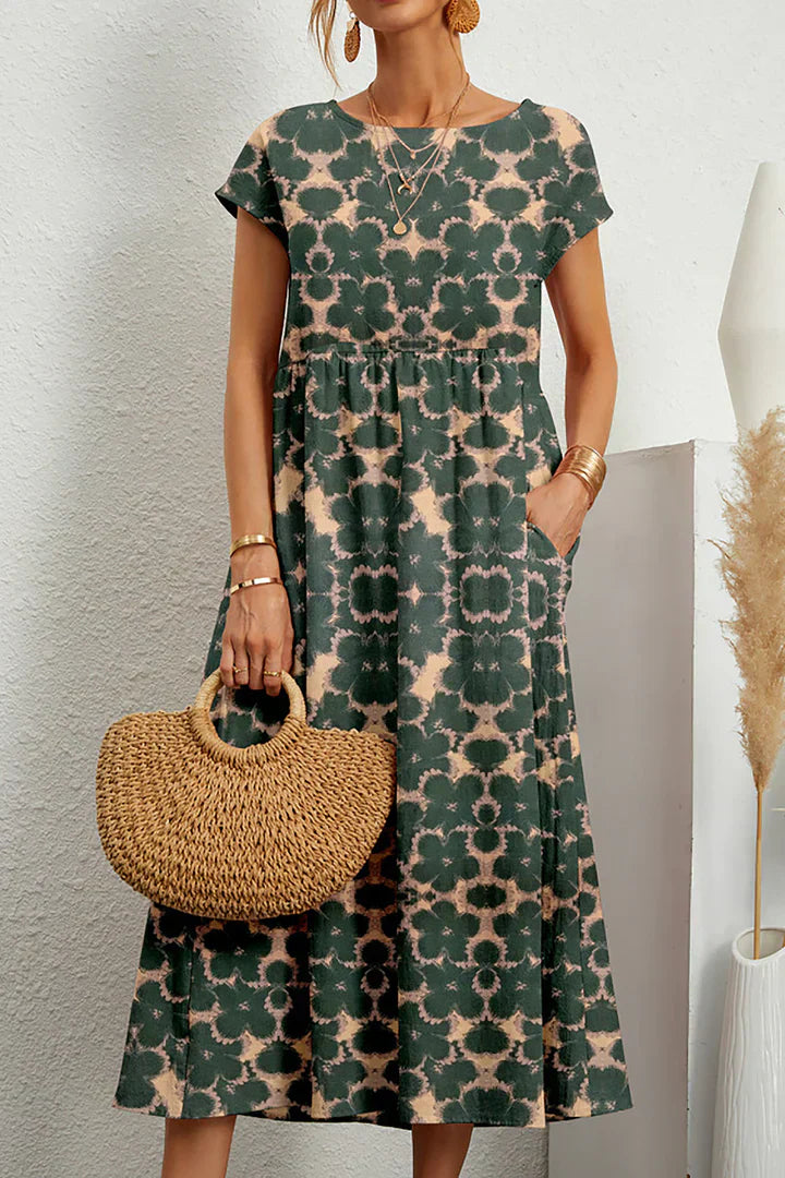 Frau in grünem Batik-Midikleid mit kurzen Ärmeln, hält runde Strohtasche, trägt goldene Accessoires. Sommermode, Boho-Stil, lässiges Outfit.