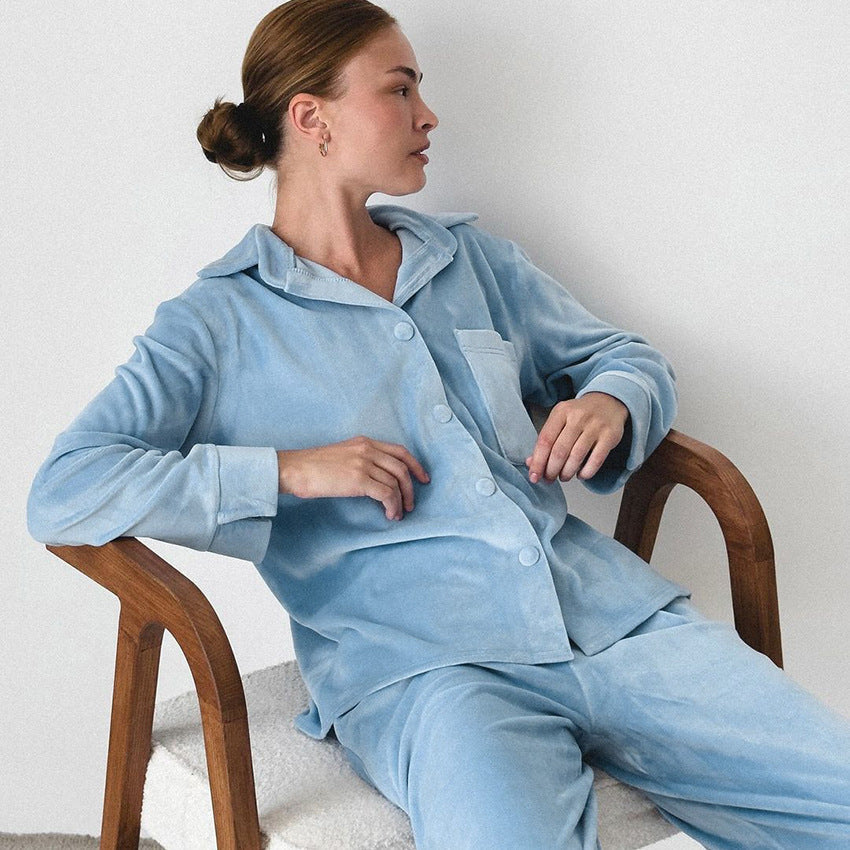 Frau in hellblauem Samt-Pyjama sitzt auf einem Holzstuhl. Bequeme Nachtwäsche, elegantes Design, entspannte Pose, minimalistischer Hintergrund.