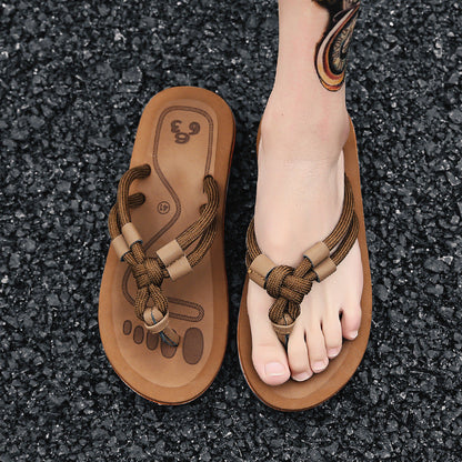 Braune Sandalen mit geflochtenen Riemen auf schwarzem Kies, Fuß mit Tattoo trägt eine Sandale. Bequeme Sommerschuhe, modisch und lässig.