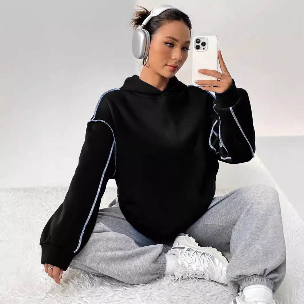 Frau mit Kopfhörern und Smartphone, trägt schwarzen Hoodie und graue Jogginghose, sitzt auf weißem Teppich. Moderne Freizeitmode, Selfie.