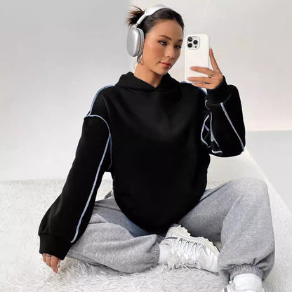 Frau mit Kopfhörern und Smartphone, trägt schwarzen Hoodie und graue Jogginghose, sitzt auf weißem Teppich. Moderne Freizeitmode, Selfie.