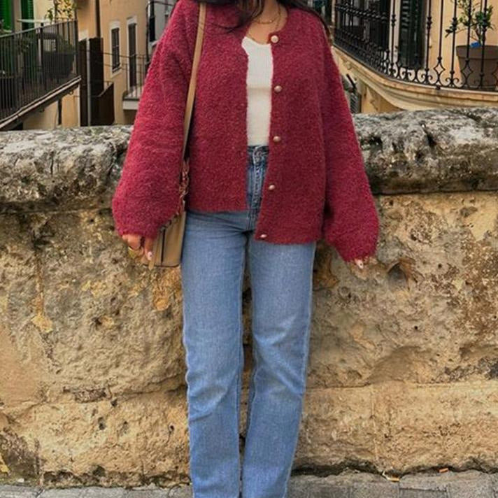 Frau in roter Strickjacke und blauer Jeans steht vor einer Steinmauer. Herbstmode, lässiger Stil, Stadtspaziergang, modische Kleidung.