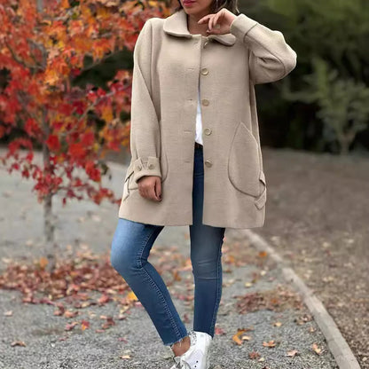 Frau in beigem Wollmantel, blaue Jeans und weißen Sneakers steht vor herbstlichem Baum. Herbstmode, Damenbekleidung, stilvoller Mantel.
