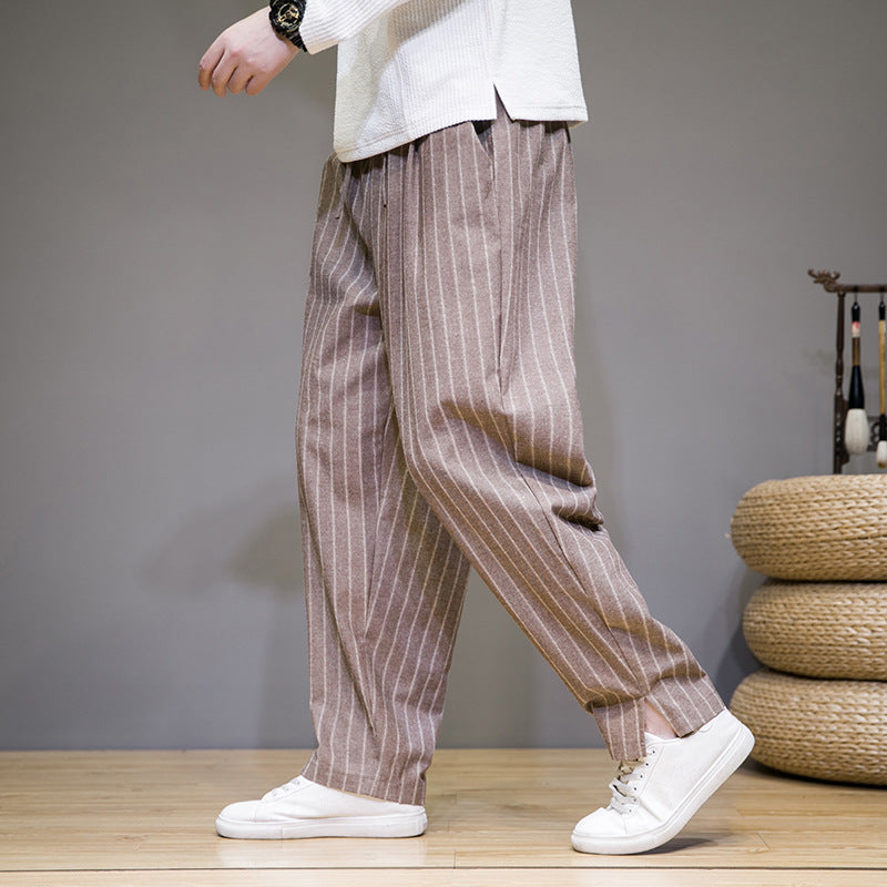 Person trägt gestreifte, beige Leinenhose und weiße Sneaker auf Holzboden. Modetrend, lässiger Stil, Herrenbekleidung, komfortable Freizeitmode.
