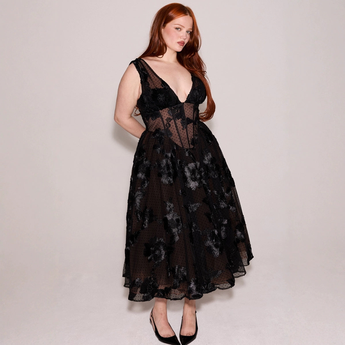 Rothaarige Frau in schwarzem, ärmellosem Spitzenkleid mit Blumenmuster, tiefem Ausschnitt und schwarzen Pumps vor neutralem Hintergrund.