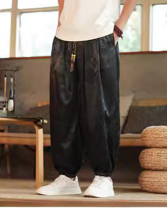 Schwarze Haremshose aus Satin, getragen von einer Person in weißem T-Shirt und weißen Sneakers, in modernem Wohnzimmer mit Holzmöbeln.