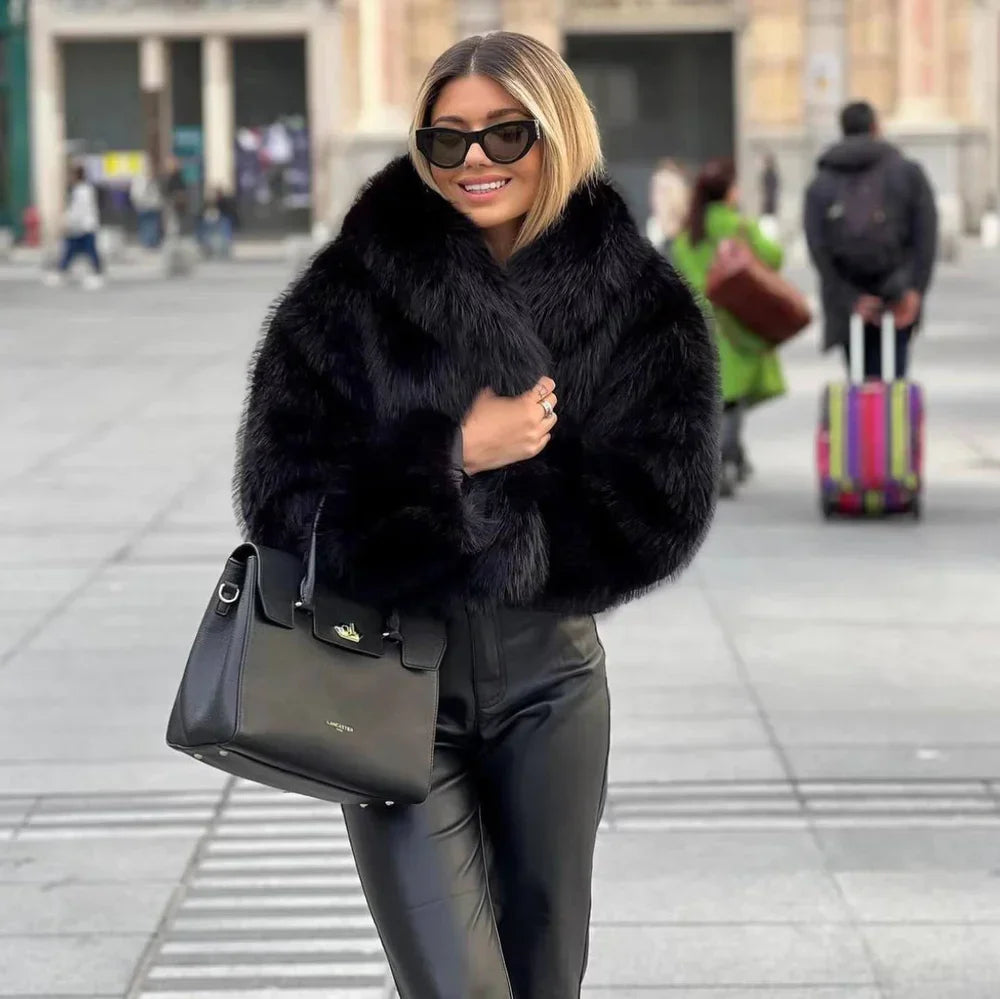 Eine Frau in schwarzem Kunstpelzmantel und Sonnenbrille, trägt eine schwarze Handtasche, steht auf einer belebten Straße. Elegante Wintermode.