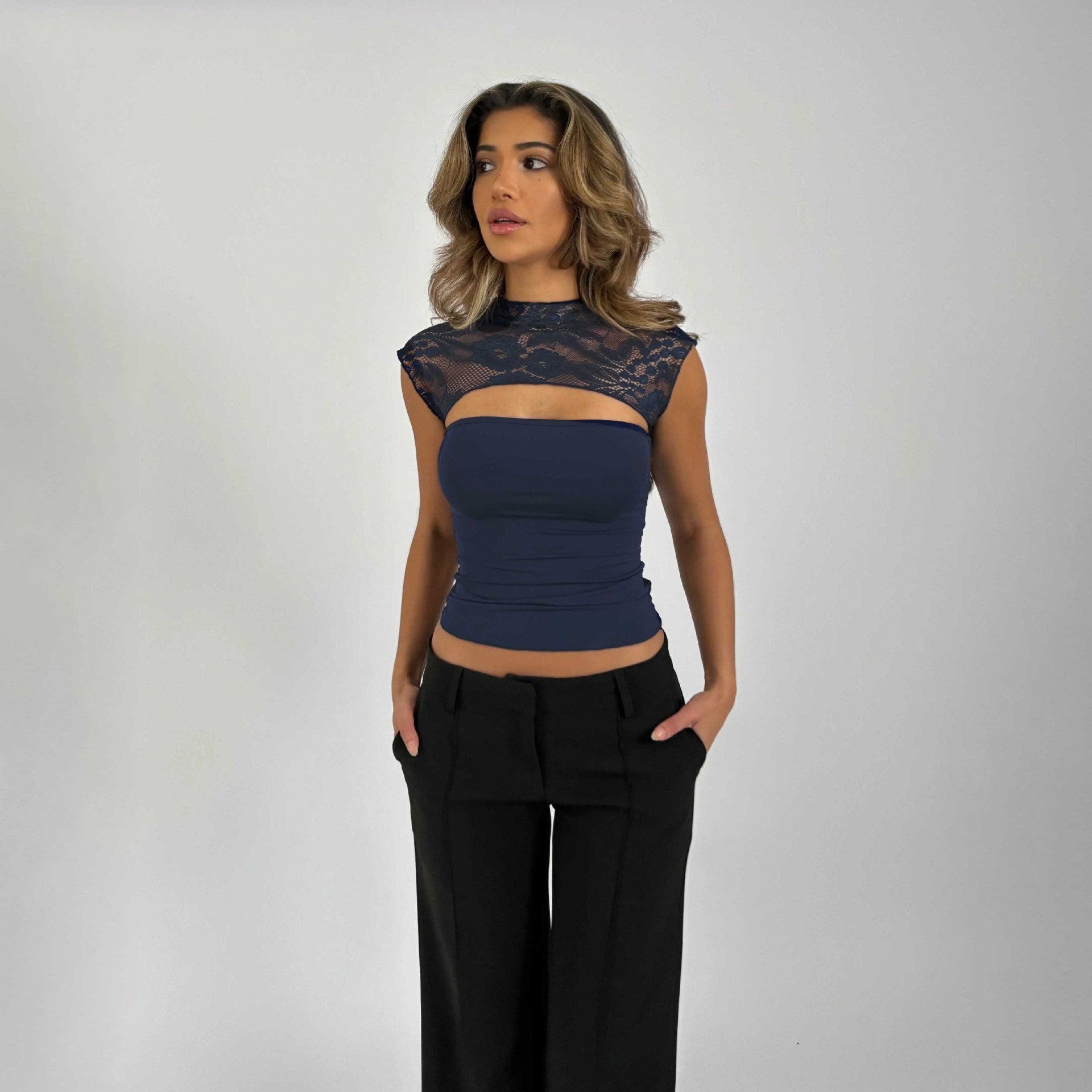 Frau in eleganter, ärmelloser Bluse mit Spitzenbesatz und schwarzer Hose vor grauem Hintergrund. Mode, Damenbekleidung, stilvolles Outfit.