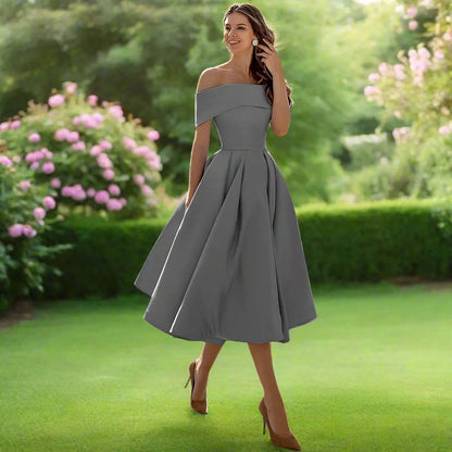 Frau in grauem, schulterfreiem Kleid im Garten, umgeben von blühenden Sträuchern. Elegantes Abendkleid, Sommermode, stilvolle Damenbekleidung.