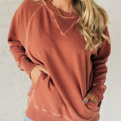 Frau in rostfarbenem Sweatshirt mit langen Ärmeln, lässiger Stil, blonde Haare, Hände in den Taschen, modische Freizeitkleidung.
