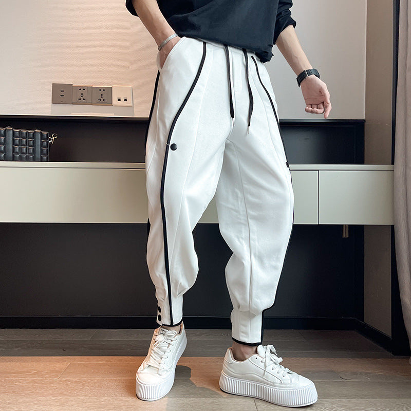 Mann trägt weiße Jogginghose mit schwarzen Streifen und weißen Sneakers, steht in modernem Raum. Trendige Herrenmode, sportlich-eleganter Look.