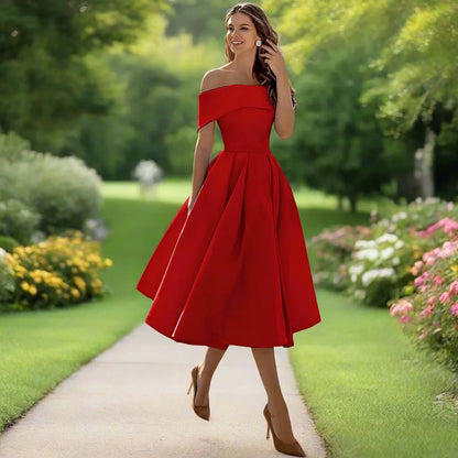 Frau in rotem, schulterfreiem Kleid spaziert auf Gartenweg, umgeben von blühenden Blumen und grünem Rasen, Sommermode, elegantes Abendkleid.