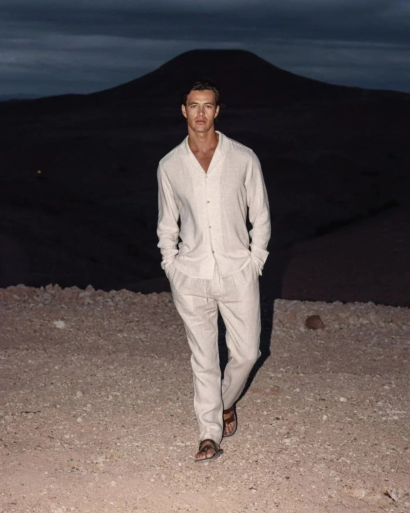 Ein Mann in einem hellen Leinenanzug und Sandalen geht in einer kargen Wüstenlandschaft bei Dämmerung. Elegante Herrenmode, Naturkulisse.