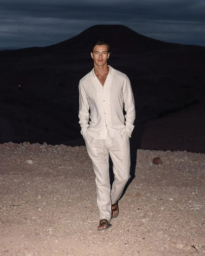 Ein Mann in einem hellen Leinenanzug und Sandalen geht in einer kargen Wüstenlandschaft bei Dämmerung. Elegante Herrenmode, Naturkulisse.