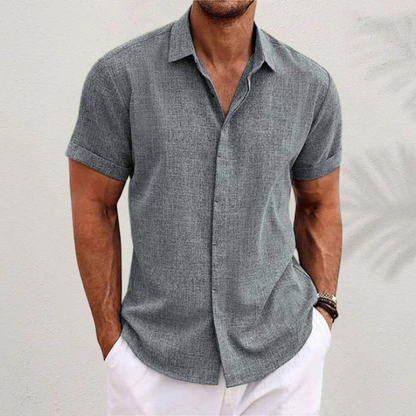 Mann in grauem Leinenhemd mit kurzen Ärmeln und weißer Hose vor neutralem Hintergrund. Sommermode, Herrenbekleidung, lässiger Stil.