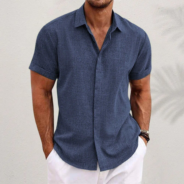 Mann in dunkelblauem Leinenhemd und weißen Shorts vor weißer Wand. Sommermode, Herrenbekleidung, Freizeitlook.