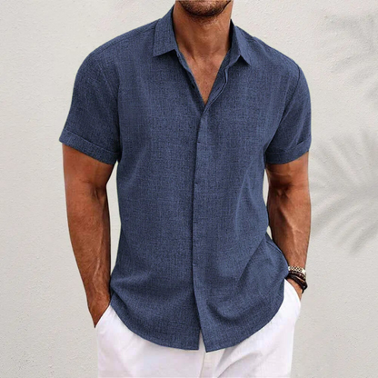 Mann in dunkelblauem Leinenhemd und weißen Shorts vor weißer Wand. Sommermode, Herrenbekleidung, Freizeitlook.
