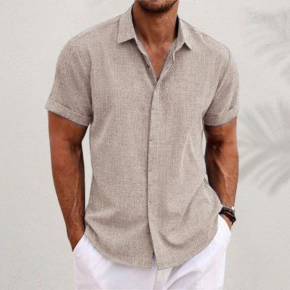 Mann in beigem Leinenhemd und weißen Shorts vor weißer Wand. Sommermode, Herrenbekleidung, lässiger Stil, kurzärmeliges Hemd.