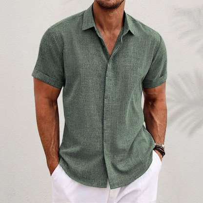 Mann in grünem Leinenhemd und weißen Shorts vor weißer Wand. Sommermode, Herrenbekleidung, lässiger Stil.