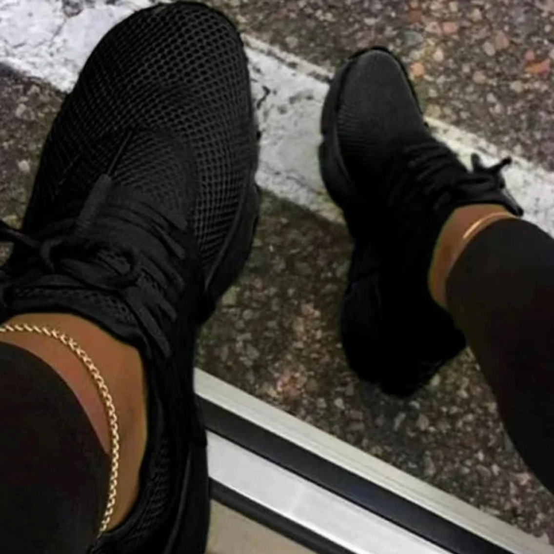 Schwarze Sneaker auf Asphalt, getragen von Person mit schwarzer Hose und goldener Knöchelkette. Modische Straßenschuhe, urbaner Stil.