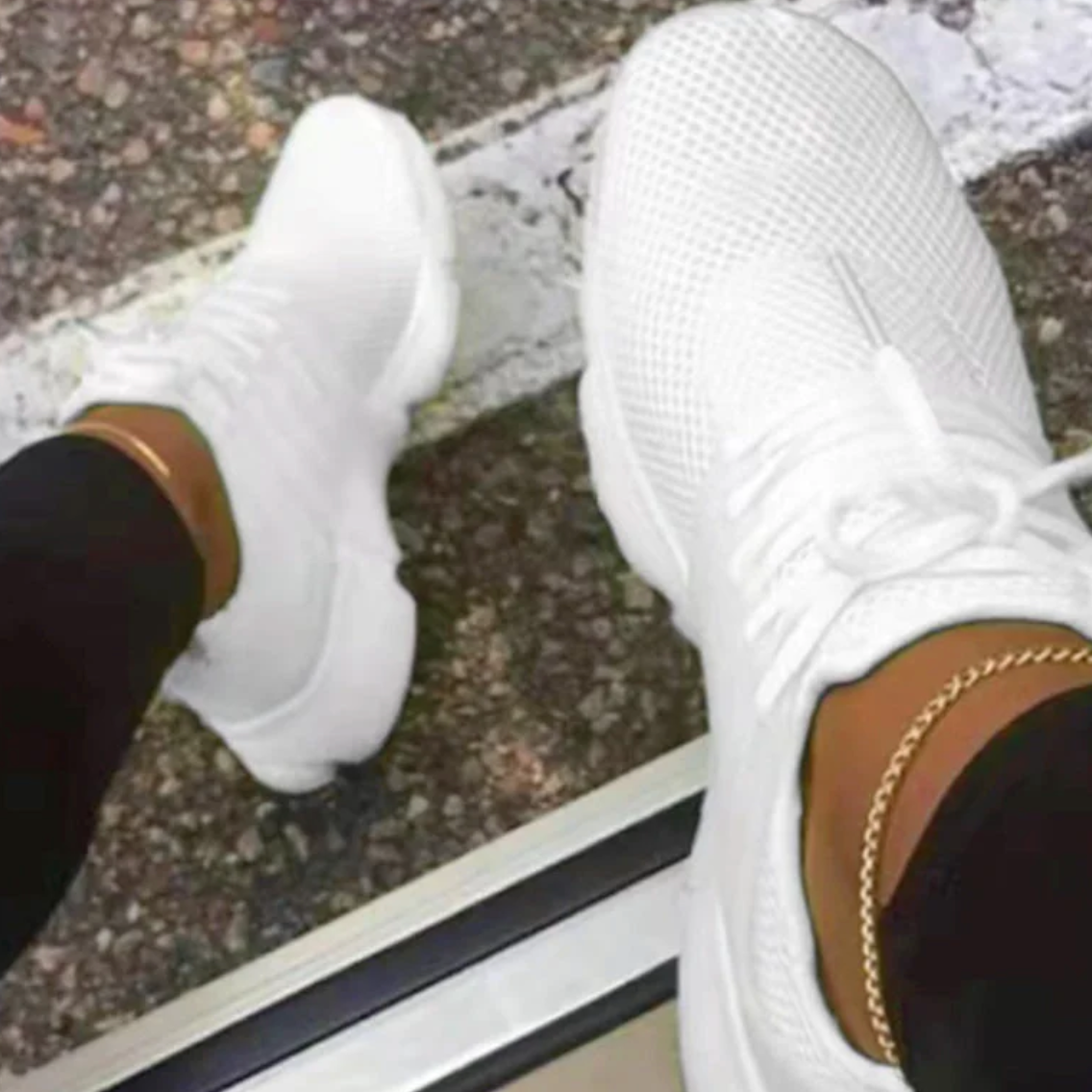 Weiße Sneaker auf Asphalt, getragen von Person mit schwarzer Hose und goldener Fußkette. Moderne Sportschuhe, urbaner Stil, Straßenmode.