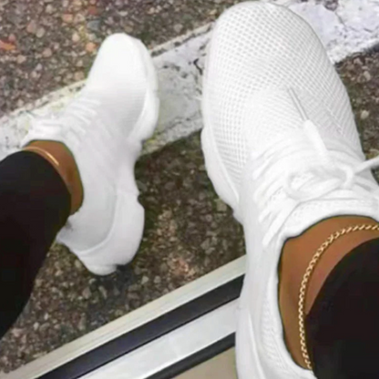 Weiße Sneaker auf Asphalt, getragen von Person mit schwarzer Hose und goldener Fußkette. Moderne Sportschuhe, urbaner Stil, Straßenmode.