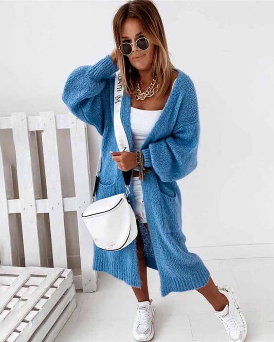 Frau in blauem Strickmantel, weißem Top und Shorts, mit weißer Umhängetasche und Sonnenbrille, steht vor weißem Hintergrund. Fashion, Streetstyle.