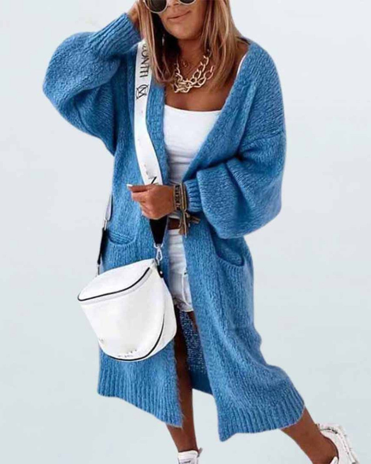 Frau in blauem Strick-Cardigan, weißem Top und Rock, mit weißer Umhängetasche und Sonnenbrille. Modetrend, lässiger Herbstlook.