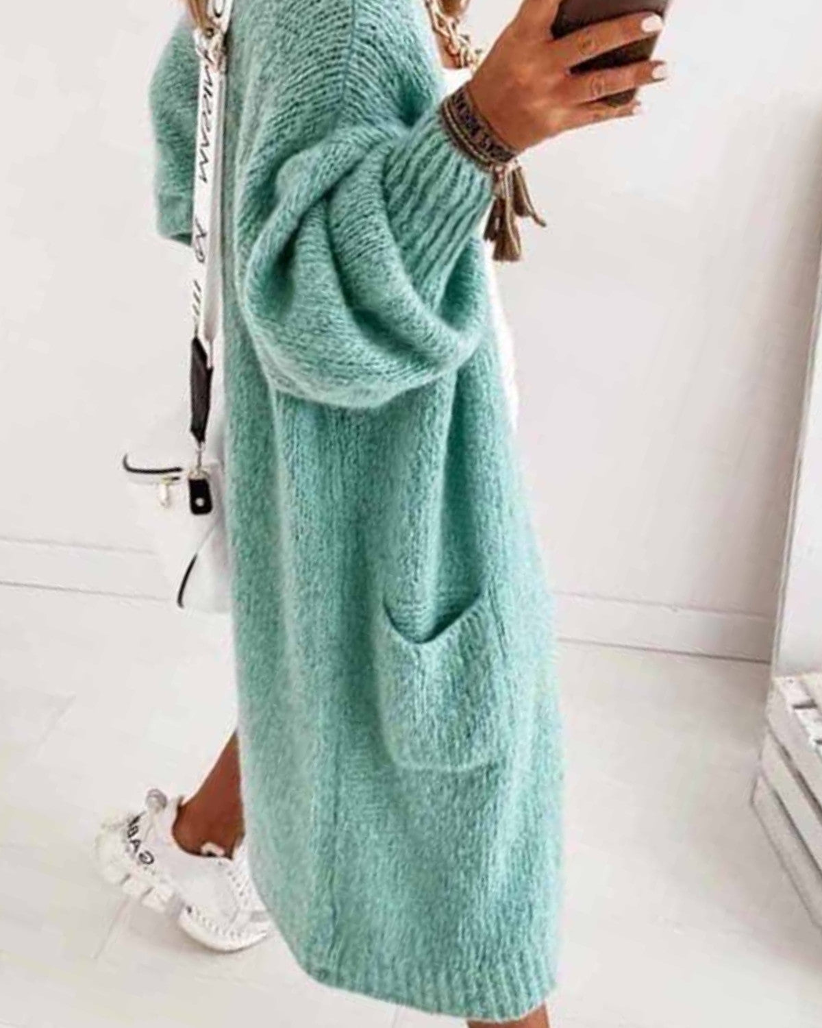 Frau in mintgrünem Strickkleid mit Taschen, hält Smartphone, trägt weiße Sneakers und weiße Umhängetasche. Modetrend, lässiger Stil.